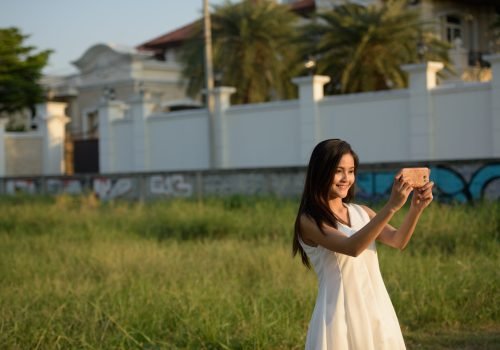 Young happy Asian woman smiling while taking selfie picture with mobile phone against grass field and fancy house
